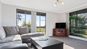 Een moderne woonkamer met een modulaire bank, een salontafel, een tv-meubel en grote ramen met uitzicht op het groen.