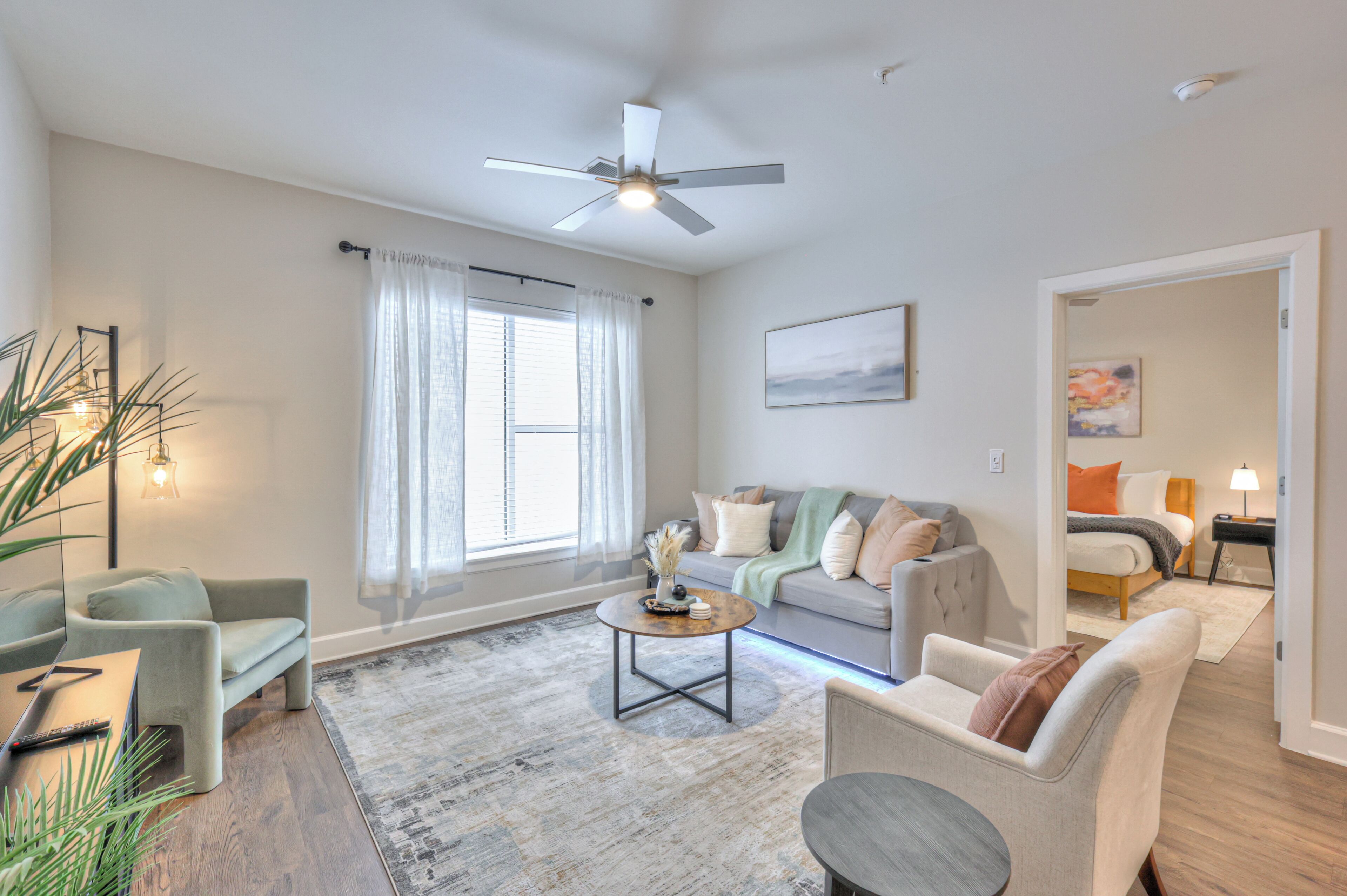 A spacious living room with a ceiling fan, a large window with curtains, a sofa set, and a coffee table.
