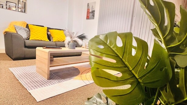 Una sala de estar moderna con un sofá gris, cojines amarillos, una mesa de centro de madera y una planta verde grande.