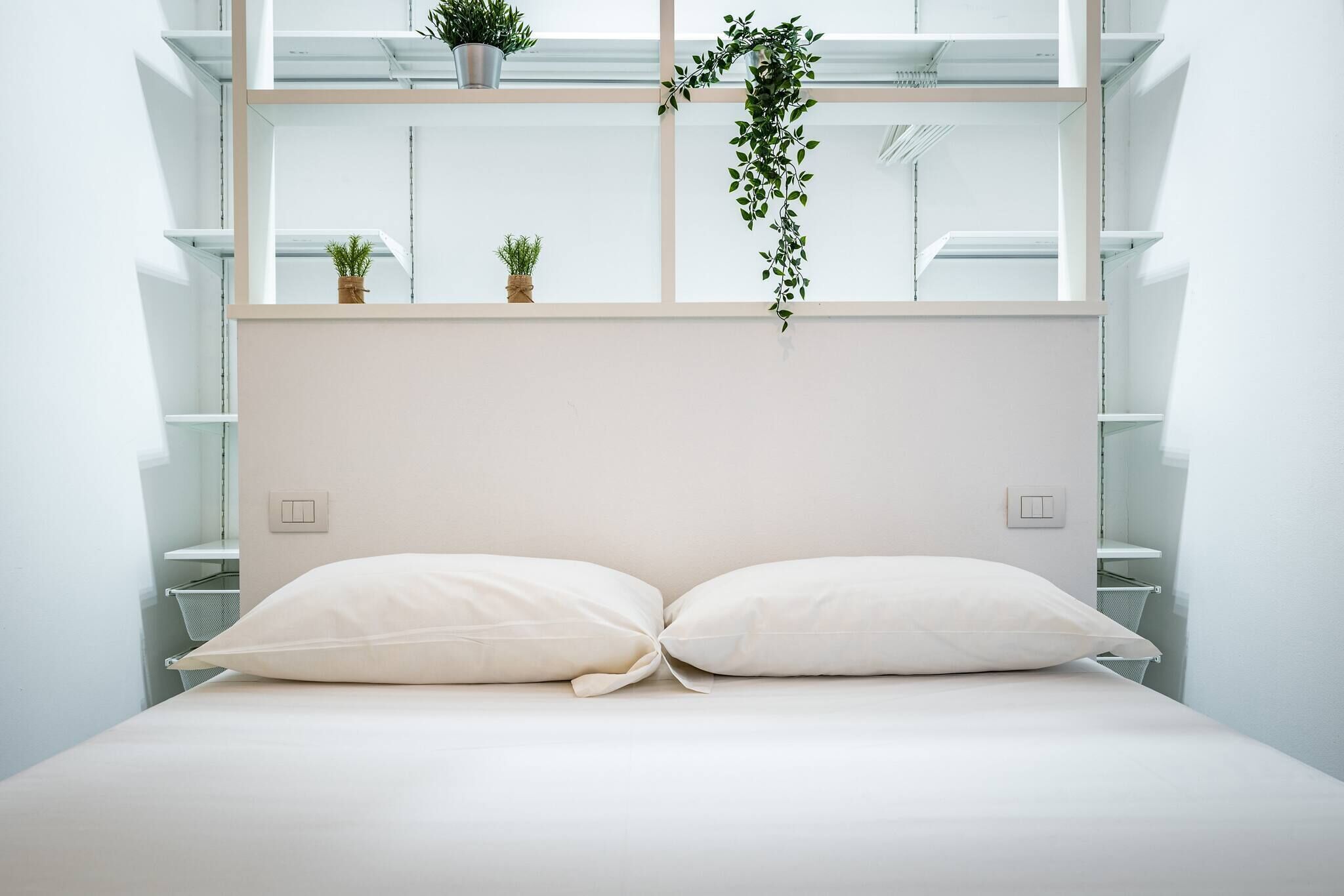 Une chambre à coucher au style minimaliste, avec un lit, deux tables de chevet, une fenêtre avec des étagères et une plante suspendue.