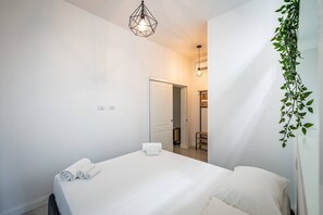 A minimalist bedroom with a bed, white linens, a hanging light fixture, and a door leading to another room.