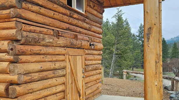 A log cabin with a wooden door and window, a concrete porch, and a wooden post.