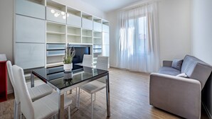 Un salon moderne avec une table à manger au plateau en verre, des chaises blanches, un canapé gris et des étagères intégrées.