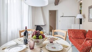 Une table à manger est dressée avec des assiettes, des couverts, un saladier rempli de fruits et une bouteille de vin. On aperçoit un canapé rouge à motifs et une lampe blanche dans la pièce.