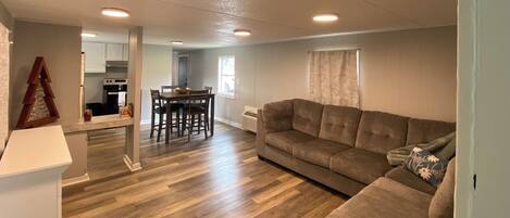 A spacious living area with a wooden floor, a brown sofa, a dining table with chairs, and a kitchen in the background.