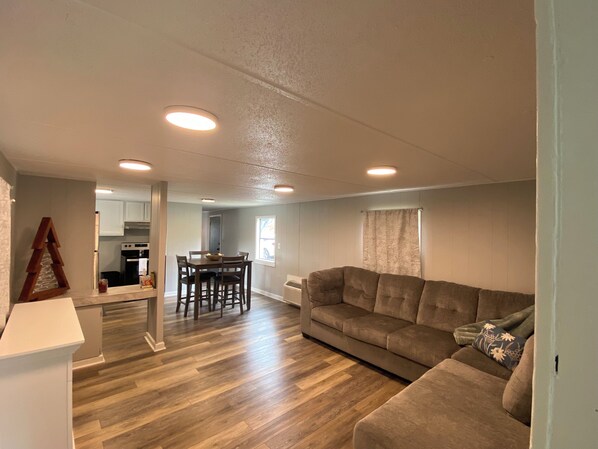 A spacious living area with a wooden floor, a brown sofa, a dining table with chairs, and a kitchen in the background.