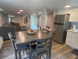 A modern kitchen with a stainless steel refrigerator, a dining table with chairs, and a gray sofa in the living area.