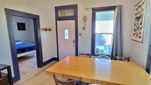 Una habitación con una mesa de comedor de madera, una cama con colcha azul, una puerta con llave y una ventana con cortinas.