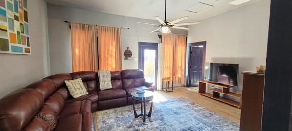 Ein Wohnzimmer mit braunem Ledersofa, einem Glastisch, einem Deckenventilator, einem Flachbildfernseher und einer Holzunterhaltungseinheit.