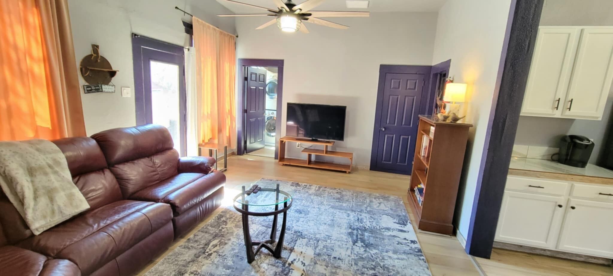 Una sala de estar con un sofá de cuero, una mesa de centro de vidrio, un televisor de pantalla plana y una zona de cocina con armarios blancos.