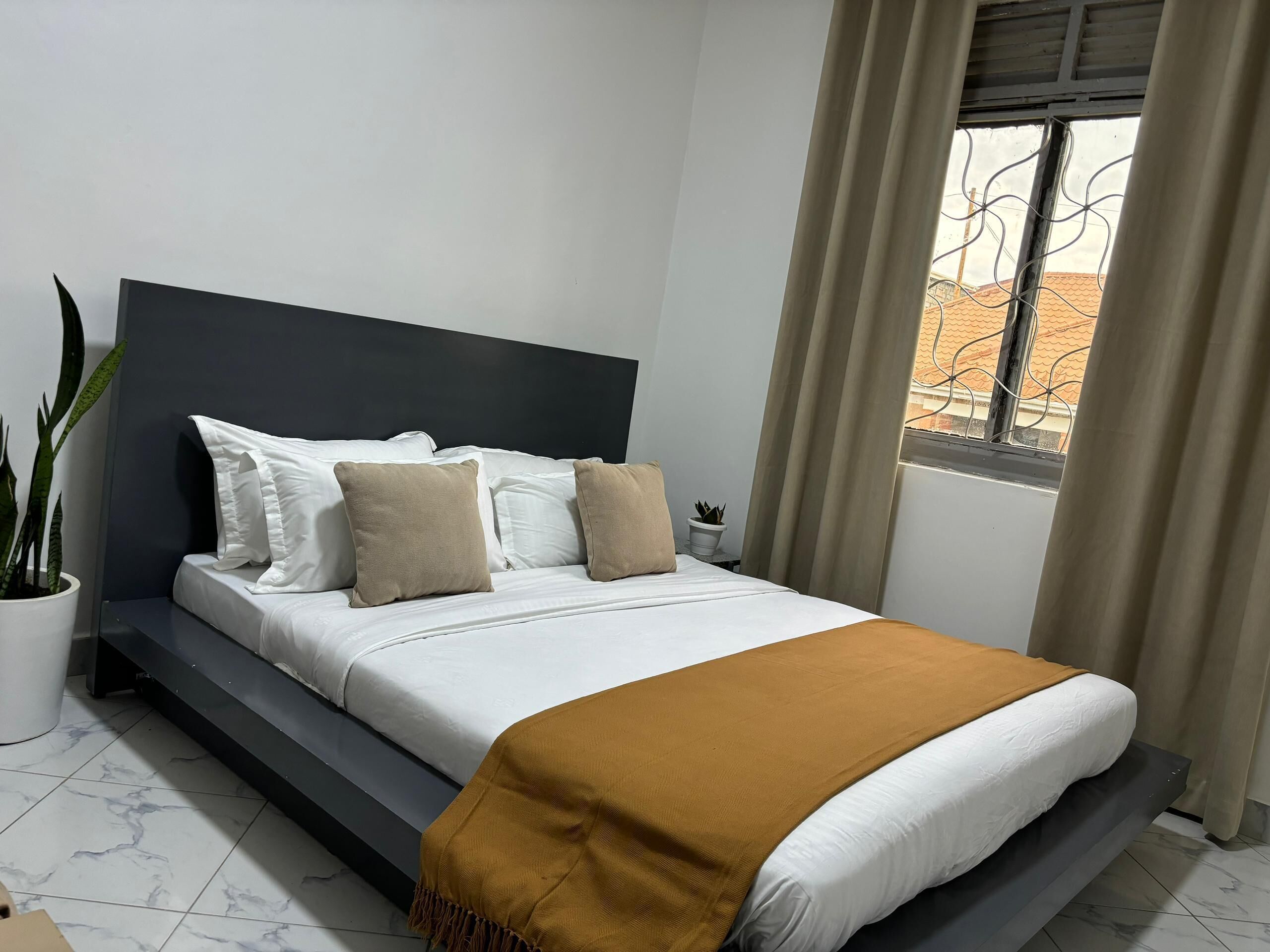 Ein Schlafzimmer mit einem Bett, einem Kopfteil, einem Fenster mit Vorhängen, einer Zimmerpflanze und einem gefliesten Boden.