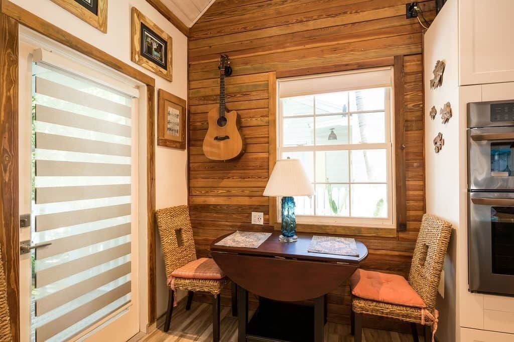 A cozy room with wooden walls, a guitar, a table with chairs, and a window with blinds.