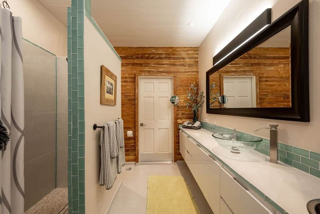 Een badkamer met een douche, een wastafel met glazen kom, een spiegel en een deur.