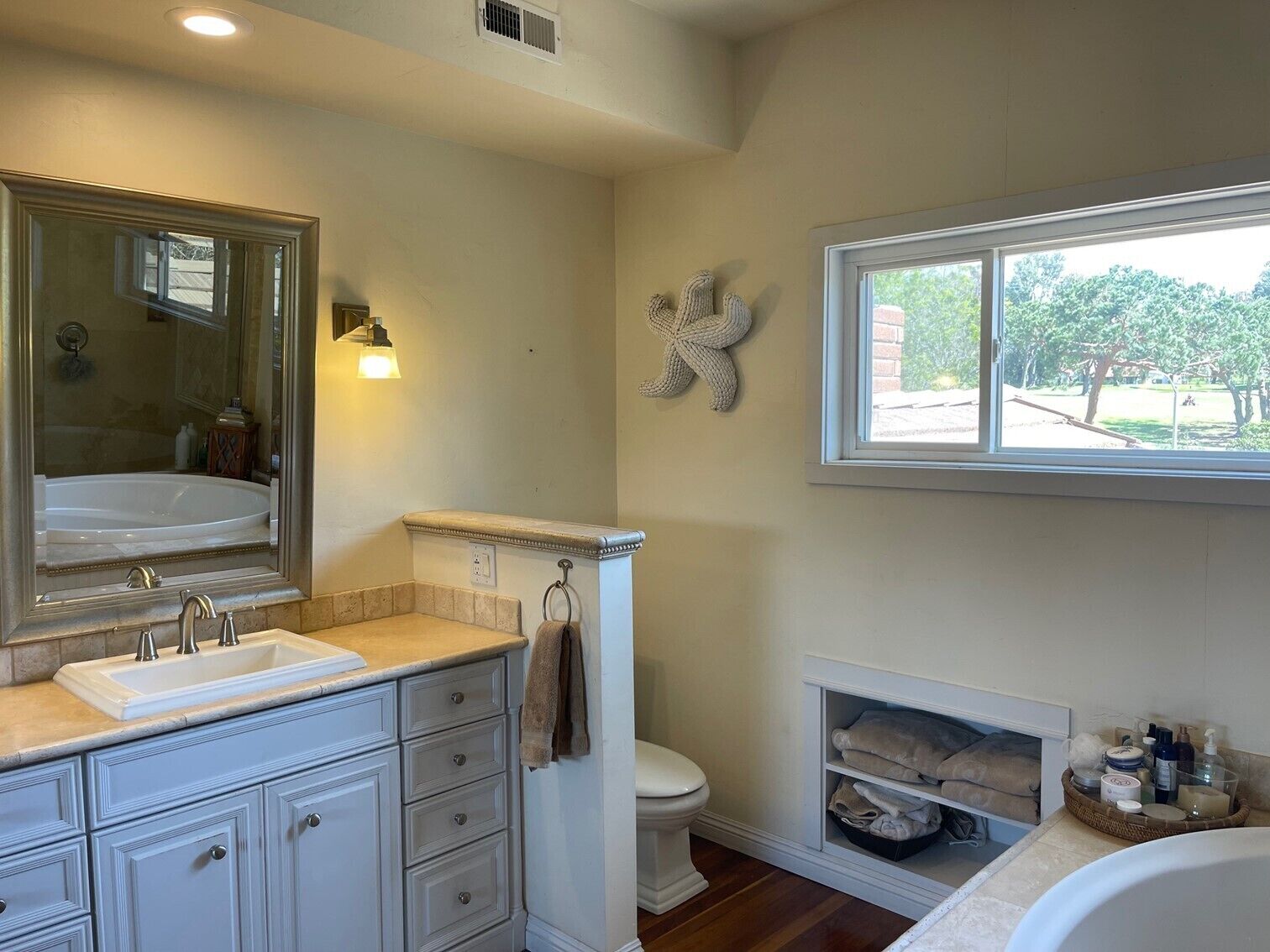 Une salle de bain avec un grand miroir, un lavabo, des toilettes et une baignoire. Une fenêtre laisse entrer la lumière naturelle et offre une vue sur l’extérieur.