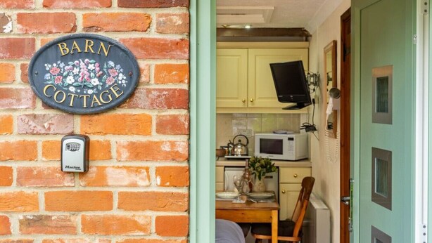 A brick wall with a 'Barn Cottage' sign and a door leading to a kitchen with a microwave and a table set for two. - Swift Host Barn Cottage Rural Barn near the Norfolk Broads (Norfolk Broads)