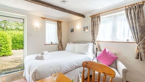 A bedroom with a bed, a wooden chair, a dining table with a glass, and a window with curtains. - Swift Host Barn Cottage Rural Barn near the Norfolk Broads (Norfolk Broads)