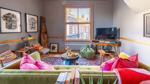 Un salon chaleureux avec un canapé vert, une table basse et un téléviseur posé sur un meuble en bois. Des tableaux encadrés ornent le mur, et une fenêtre offre une vue sur les bâtiments environnants.