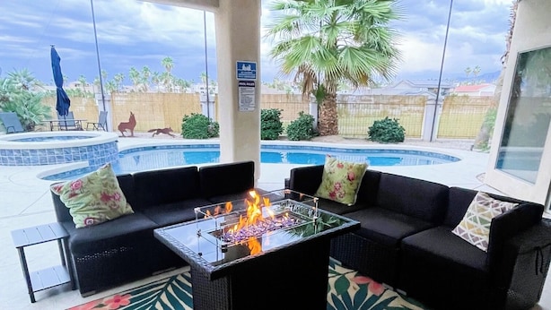Un salon au bord de la piscine avec une table à plateau en verre, des canapés noirs et un tapis à motifs floraux.