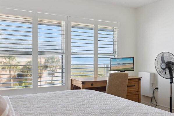 A bedroom with a bed, a desk with a computer, a standing fan, and a window with blinds.