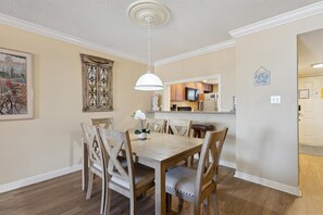 A dining area with a wooden table and chairs, a hanging light fixture, and a kitchen visible through an open doorway.