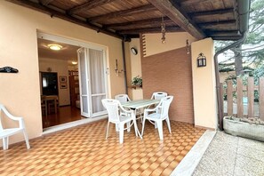 Una veranda esterna con pavimento in legno, sedie bianche e un tavolo.