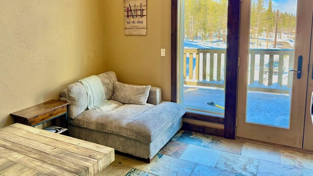 Una habitación acogedora con un sofá a rayas, una mesa de centro de madera, una lámpara de pared y un amplio ventanal con vistas a una terraza nevada y árboles.