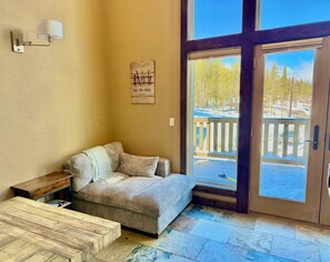 A cozy room with a striped sofa, a wooden coffee table, a wall-mounted lamp, and a large window with a view of a snowy deck and trees. - Spacious town house in Kirkwood (KIRKWOOD)