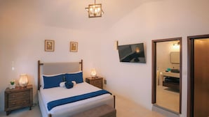 Ein modernes Schlafzimmer mit Bett, Nachttischen, einer Bank, einem Fernseher und einem durch eine offene Tür einsehbaren Badezimmer.