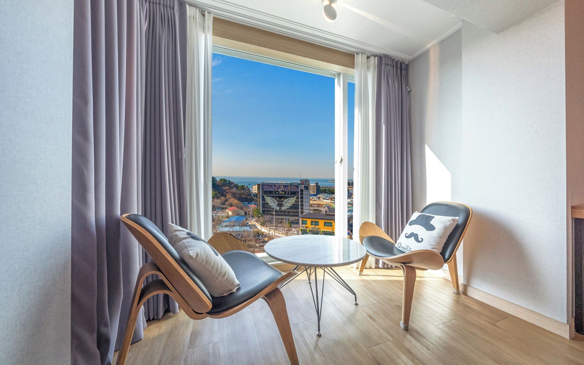 A modern room with two chairs and a small table, large windows with curtains, and a view of the cityscape.