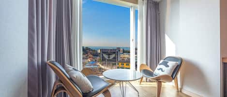 A modern room with two chairs and a small table, large windows with curtains, and a view of the cityscape.