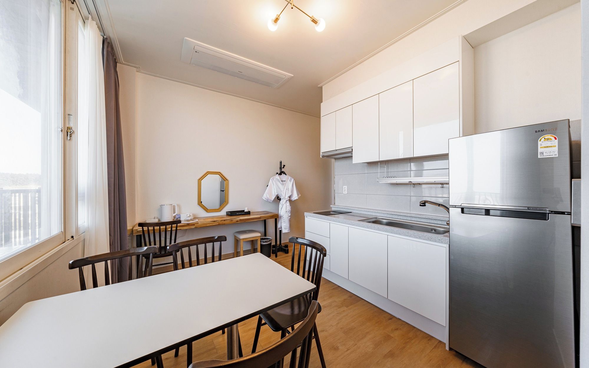 A compact kitchen with a stainless steel refrigerator, a dining table with chairs, and a window with curtains.