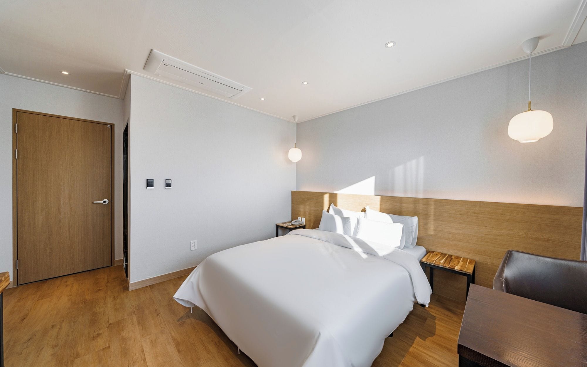 A modern hotel room with a large bed, bedside tables, a wooden headboard, and a desk with a chair.