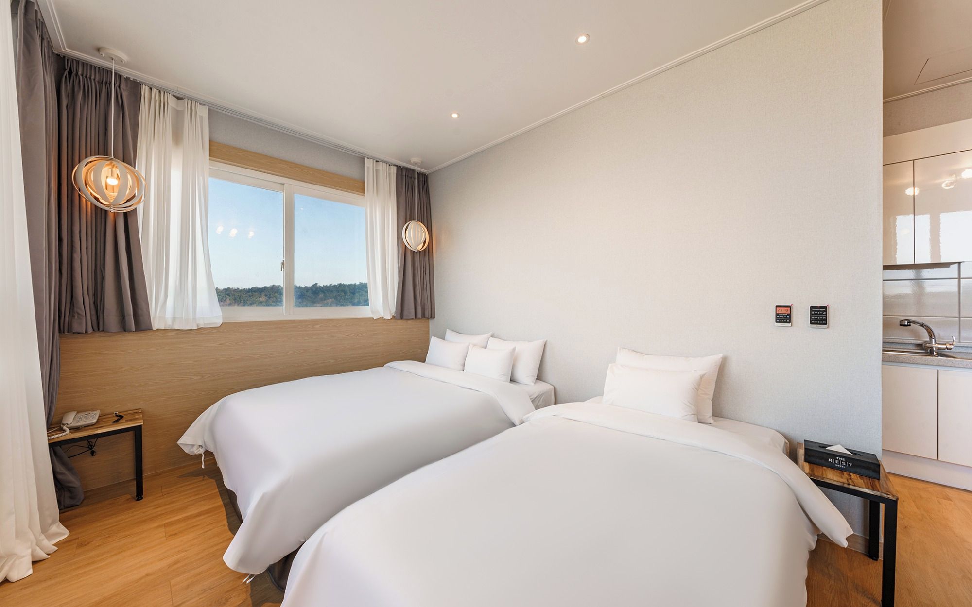 Een hotelkamer met twee bedden, een houten hoofdeinde, een raam met gordijnen, een nachtkastje met een telefoon en een klein bureau met een lamp.