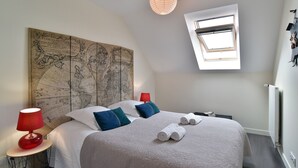 Et soveværelse med et stort kort på væggen, ovenlys, en seng med hvidt sengetøj og blå puder samt en rød lampe på et sengebord.
