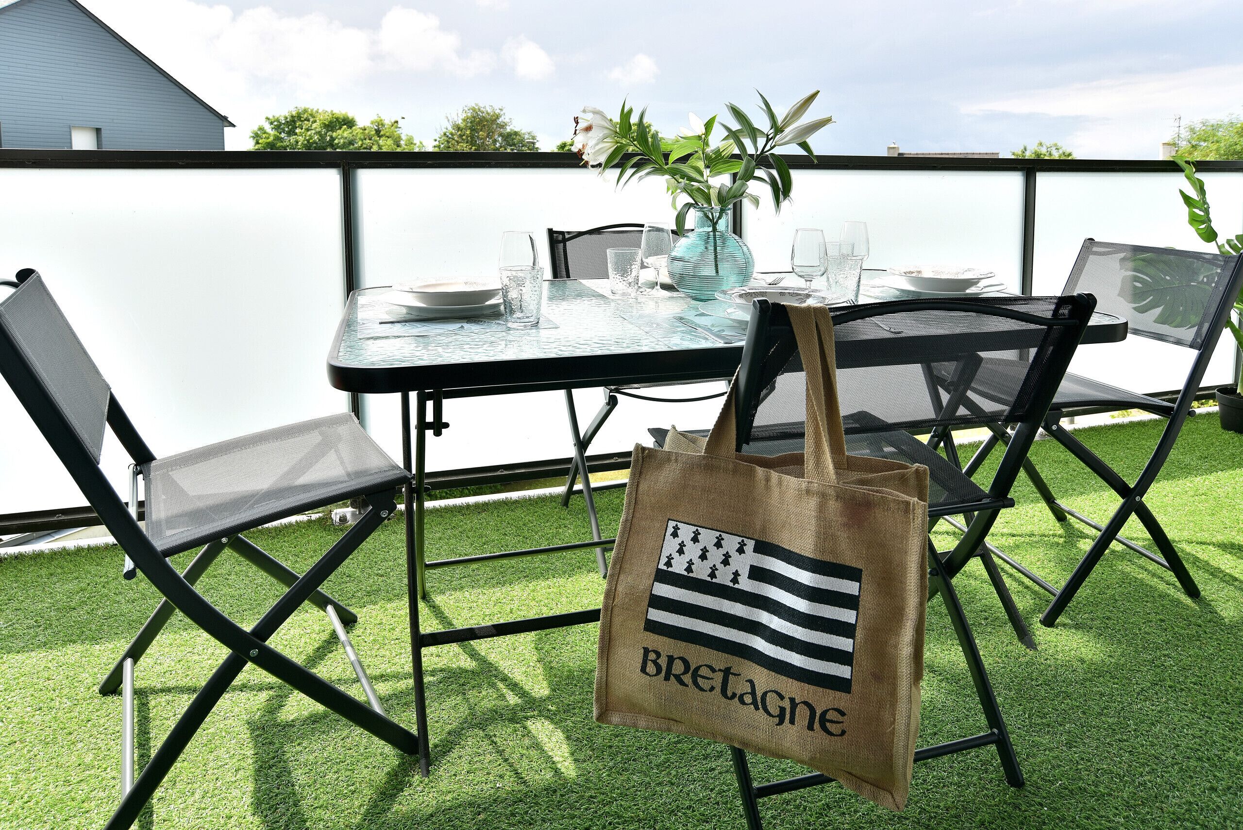 An outdoor dining area with a table set for two, chairs, a vase with flowers, and a tote bag with a flag design.