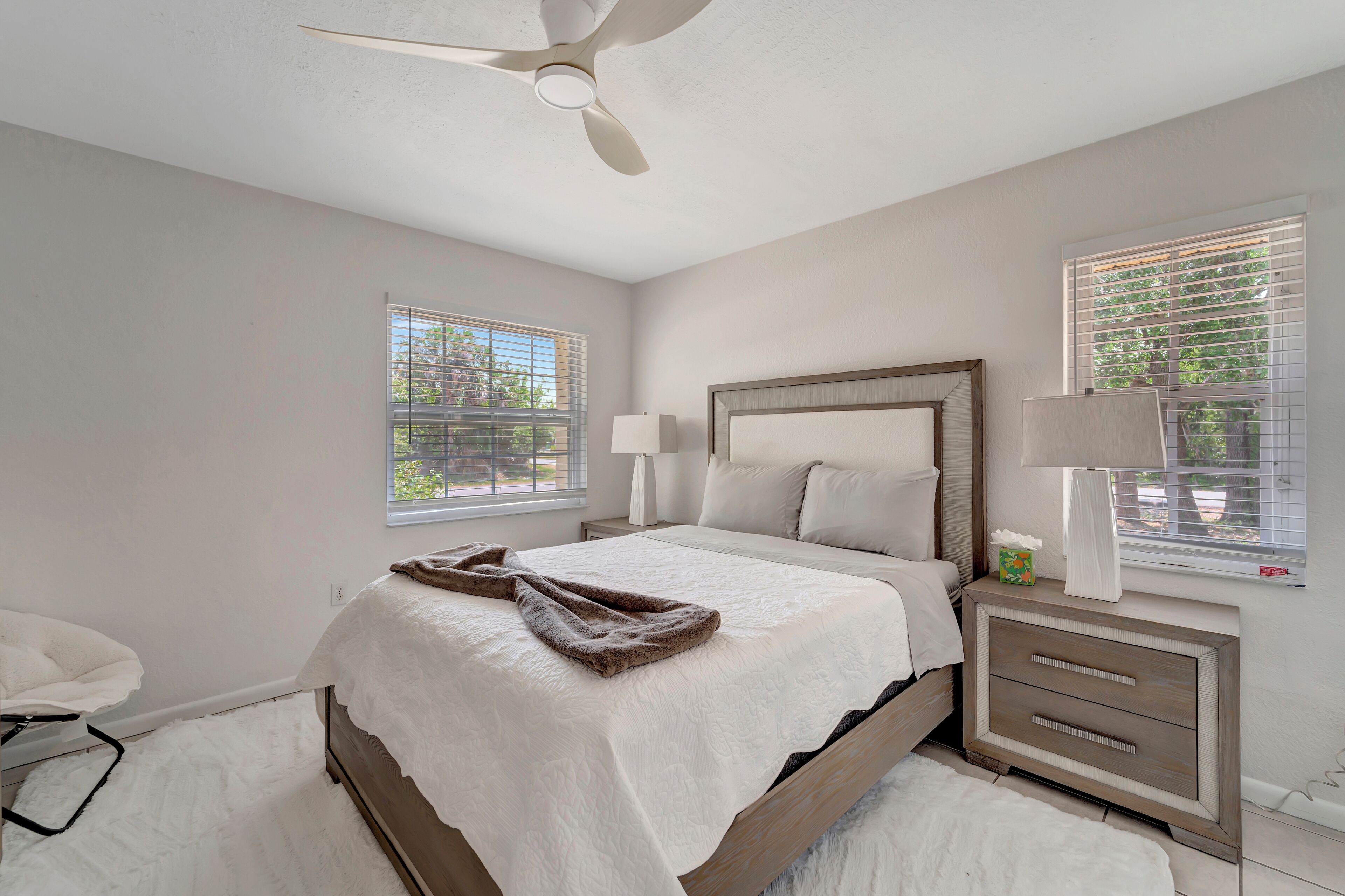 A bedroom with a bed, bedside tables, a ceiling fan, and windows with blinds.