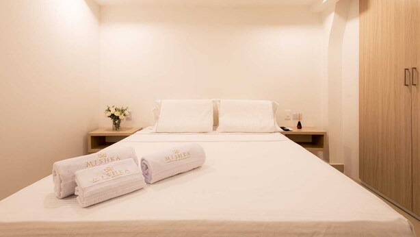 Quarto de hotel com cama bem arrumada, duas mesas de cabeceira com um vaso de flores e um guarda-roupa de madeira.