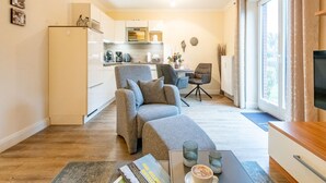 Un salon moderne avec un canapé, une table basse et un téléviseur. On aperçoit une cuisine au fond, avec un four à micro-ondes et une bouilloire.