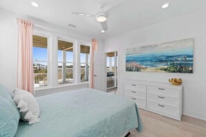 A bedroom with a bed, a ceiling fan, a painting, and a view of the outside. - At The Pointe by Brunswick Vacation Rentals: 4BR 4.5BA w/ Pool, Spa, EV Charger (Ocean Isle Beach)