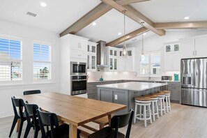 A modern kitchen with white cabinetry, stainless steel appliances, a wooden dining table, and bar stools. - At The Pointe by Brunswick Vacation Rentals: 4BR 4.5BA w/ Pool, Spa, EV Charger (Ocean Isle Beach)