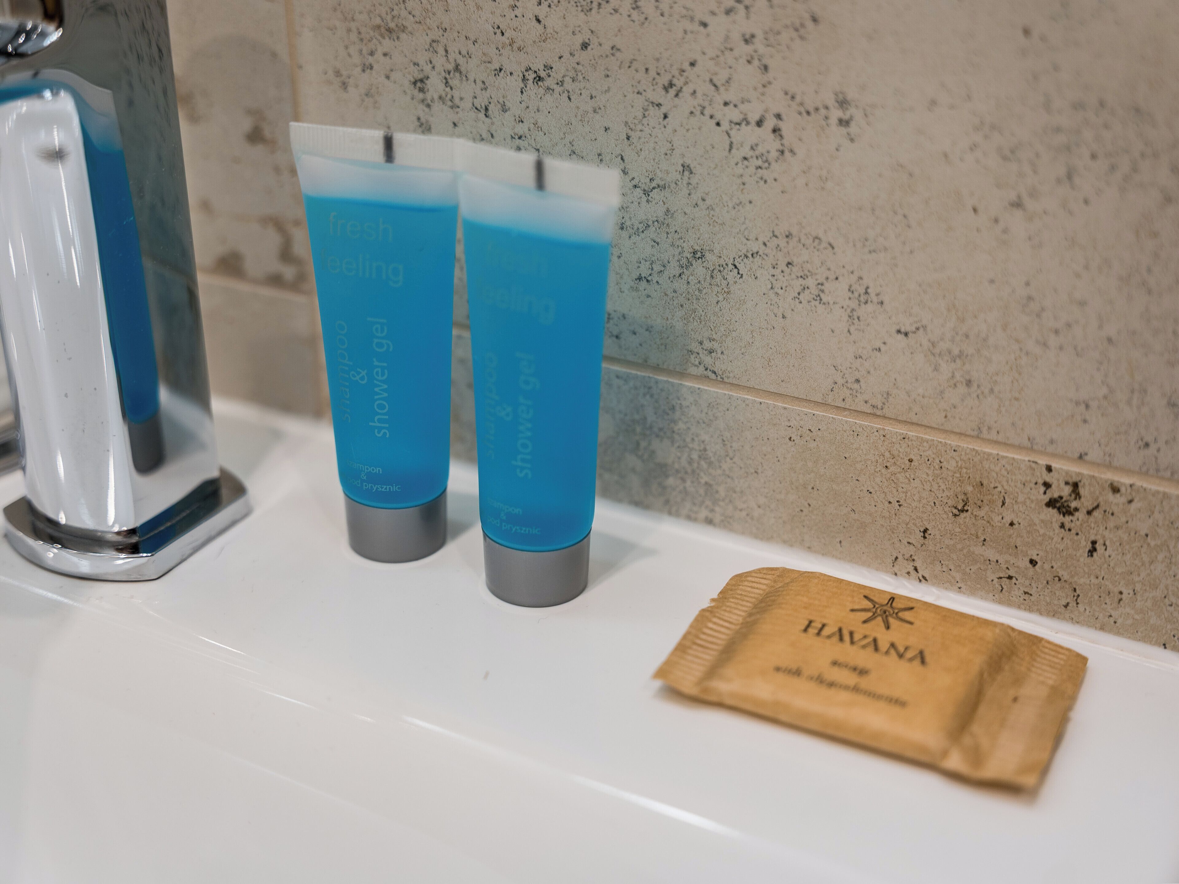 Un baño con un lavamanos blanco, dos botellas azules de champú y gel de ducha, y una bolsa marrón con la etiqueta "HAVANA".