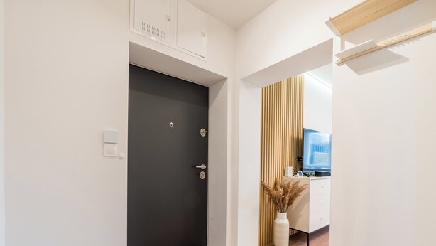 A hallway with a dark door, a wooden floor, a white wall, and a wooden panel on the right.