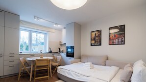 A modern, well-lit room with a kitchenette, a dining area with a round table and chairs, a large bed with pillows, and two framed pictures on the wall.
