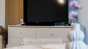 A modern living room with a large flat-screen TV on a stand, a white cabinet below, and a vase with flowers on a table.