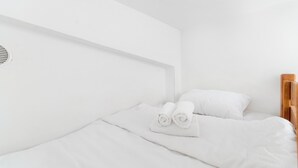 A single bed with white bedding and pillows, a wooden headboard, and a white wall with a vent.