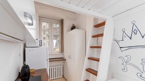 A compact kitchen with a microwave, a small refrigerator, and a wooden table. A white radiator is visible, and a window allows natural light. A white cabinet with a crown design is on the wall.