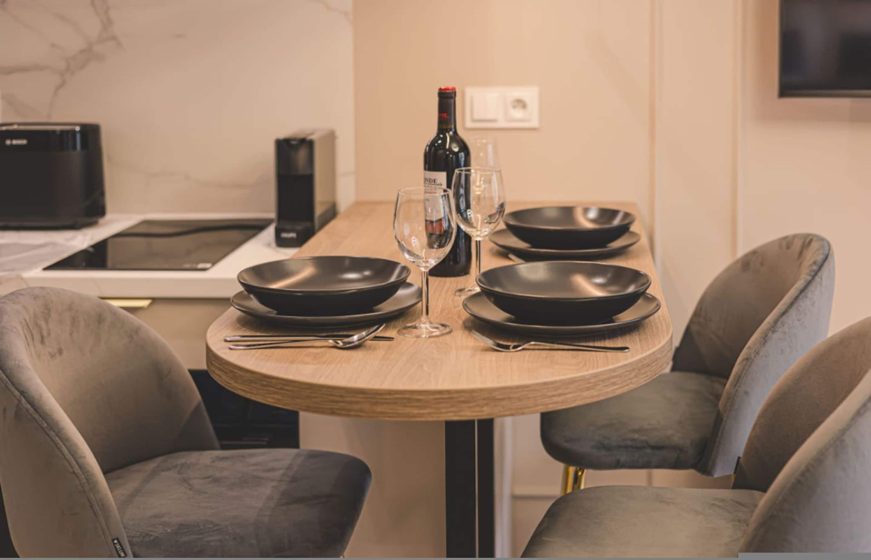 Een eettafel met vier zwarte borden, twee wijnglazen en een fles wijn. Rond de tafel staan vier stoelen bekleed met grijs stof. In de achtergrond is een zwarte magnetron en een witte inductiekookplaat zichtbaar.
