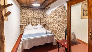 Un dormitori amb paret de pedra, bigues de fusta al sostre, un llit amb edredó blanc, una cadira de fusta i una taula petita.
