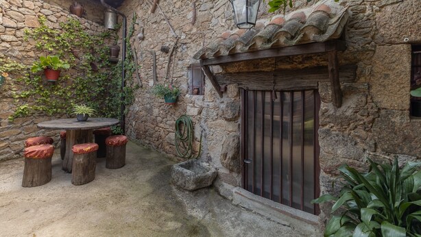 Edifici de pedra rústic amb teulada de teules, una porta de fusta i una taula rodona amb tamborets.