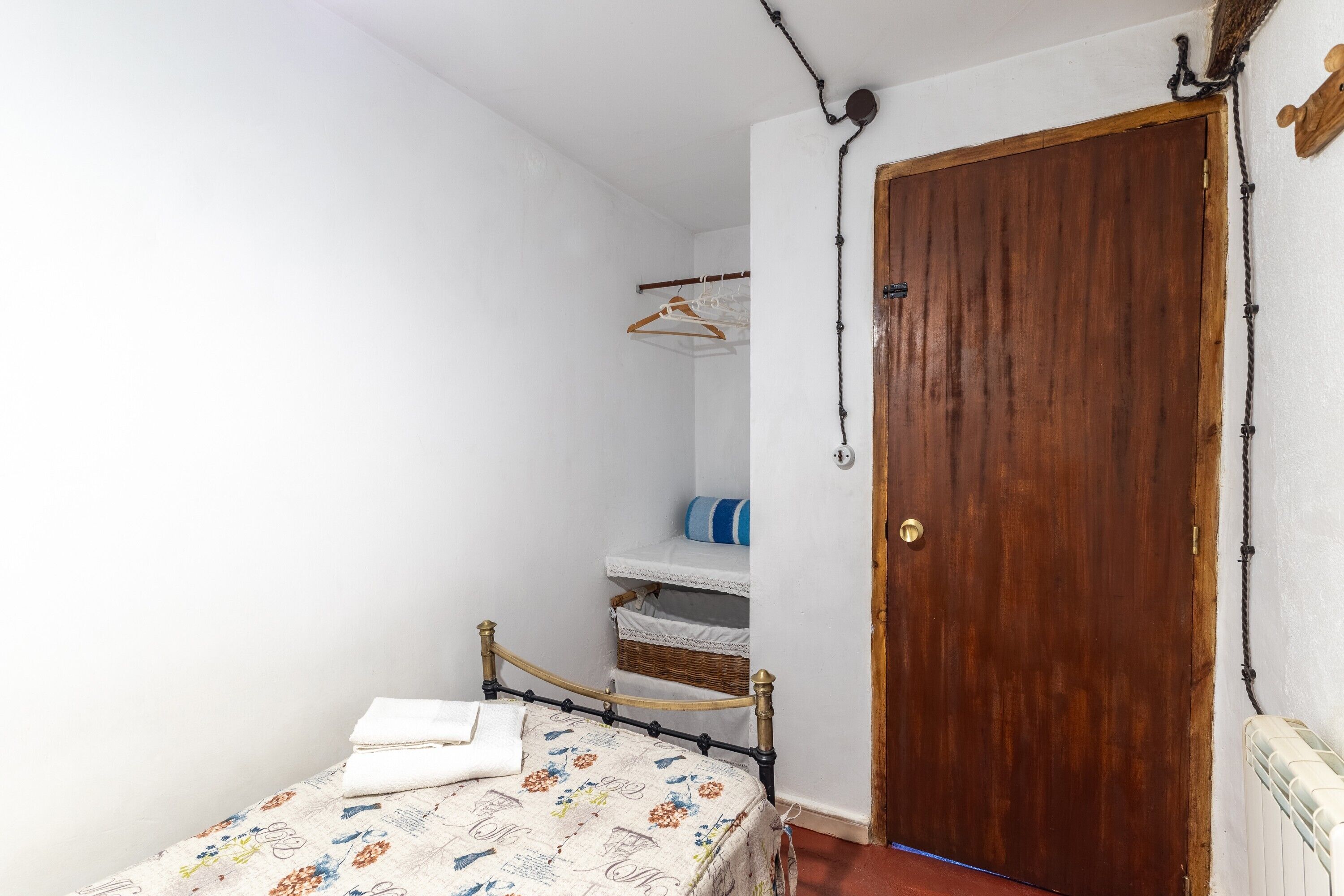 Une chambre avec un lit simple, une porte en bois, un mur blanc et une petite étagère sur laquelle est suspendu un vêtement.